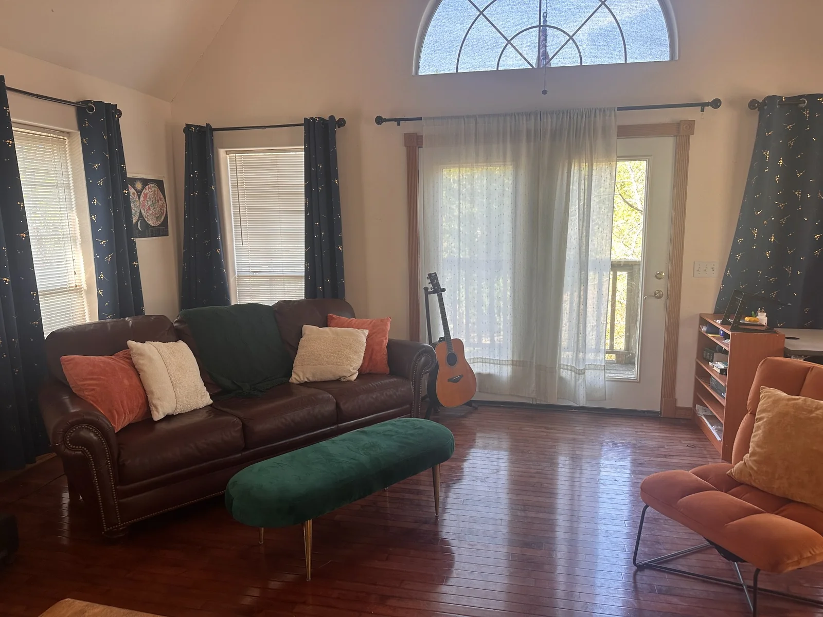 Conservatory seating area with a brown sofa, green ottoman, guitar, and tall arched window